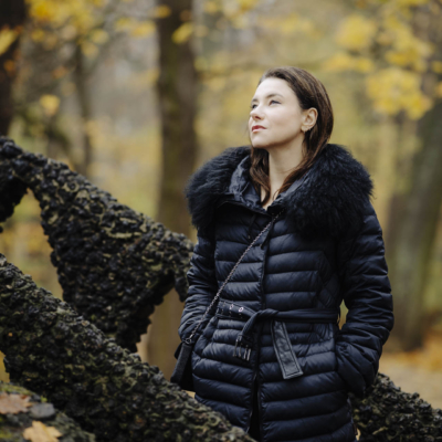 Anna w zimowym stroju, stojąca na tle jesiennych drzew w toruńskim parku, w nastrojowym świetle popołudnia.
